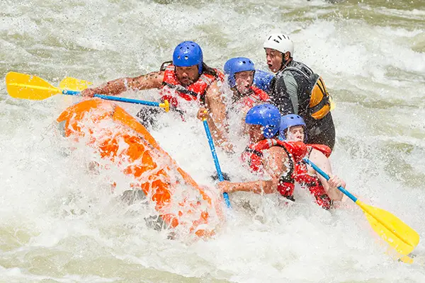 Group of people wearing helmets and life jackets paddling an orange raft through splashing whitewater rapids with turbulent water surrounding them conveying excitement and teamwork in a fast-moving river environment