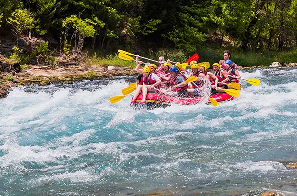 Group of people wearing helmets and life jackets paddling a raft through fast-moving whitewater rapids surrounded by lush green trees and rocky riverbanks conveying excitement and adventure