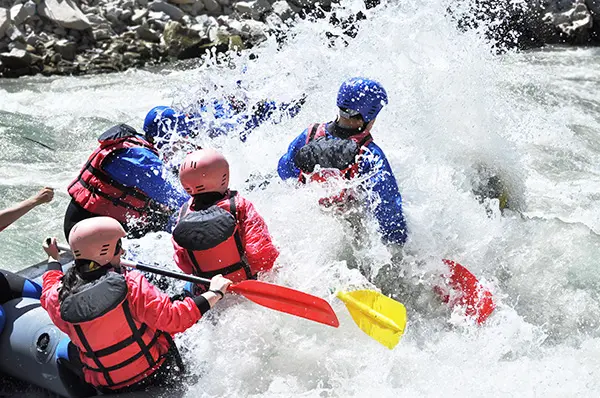 Group of people wearing helmets and life jackets paddling a raft through splashing whitewater rapids with rocky riverbanks in the background conveying excitement and teamwork