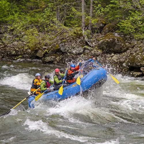 Rafting boat with several people paddling through fast whitewater rapids surrounded by splashing waves and rocky riverbanks under a bright sky conveying excitement and adventure