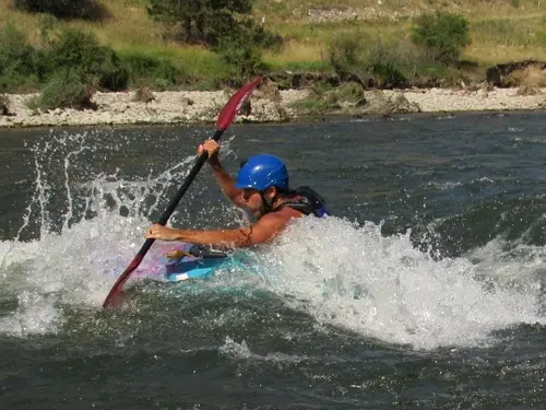 Person wearing a blue helmet and life jacket paddling a kayak through splashing whitewater rapids on a fast-moving river surrounded by green trees and rocky riverbank conveying excitement and adventure